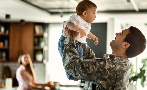 Happy army soldier playing with his baby son and having fun at home. Mother is in the background.