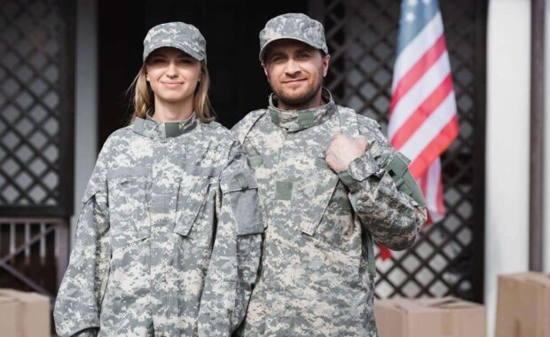 smiling military couple looking at camera near cardboard boxes, with blurred house on background